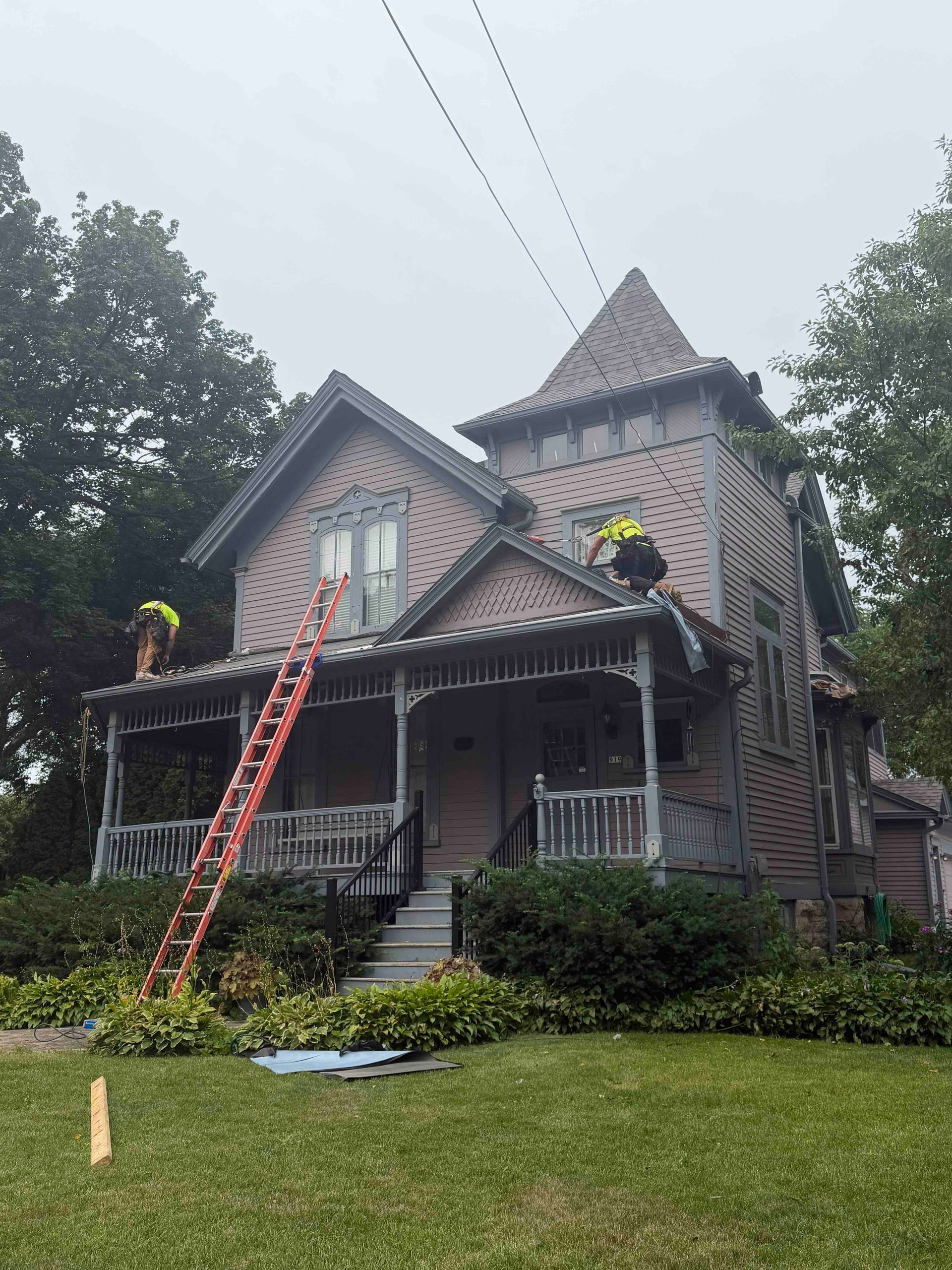 Victorian house with porch and roof under repair, ladders leaning on the front, grassy yard in front