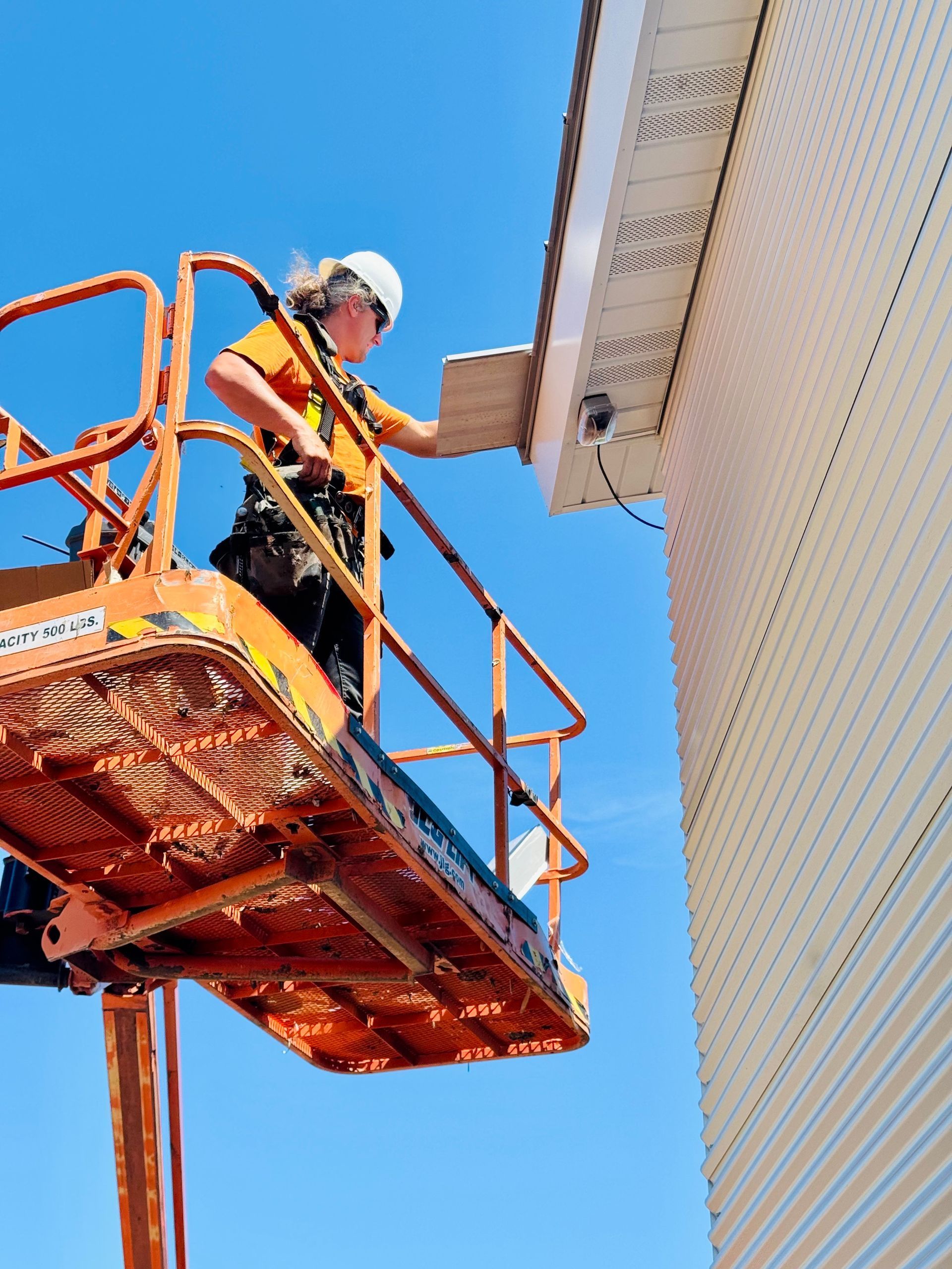 Worker on an orange lift inspecting the underside of a roof against a blue sky