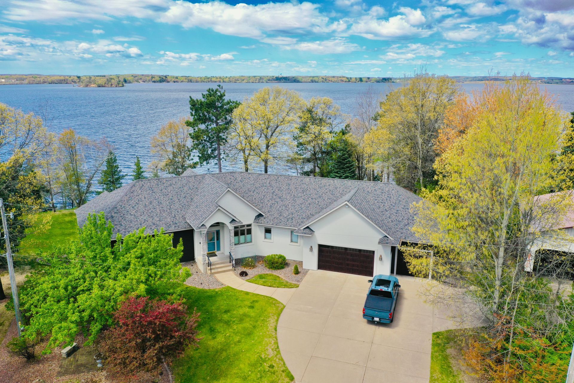 Aerial view of a lakeside house with a driveway, green lawn, and autumn trees.