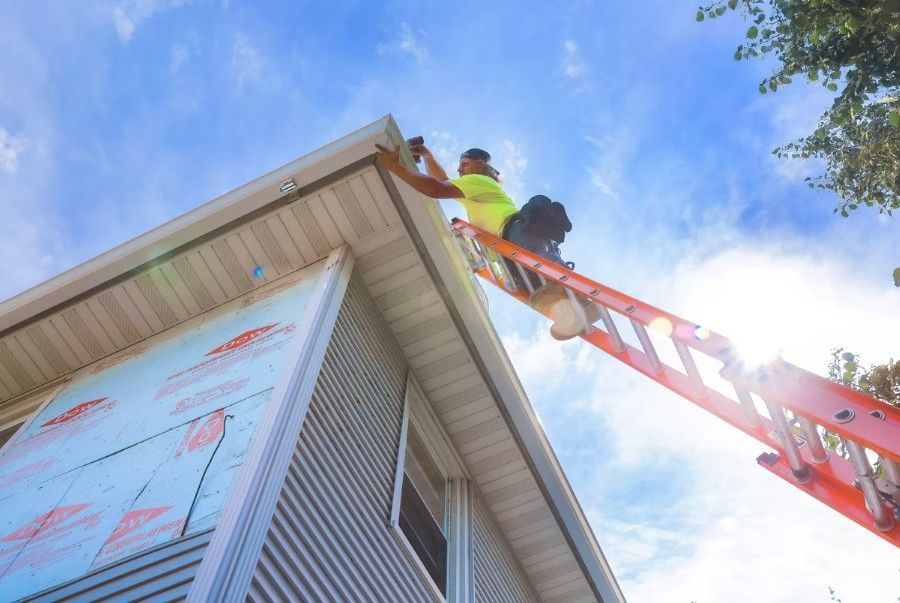 Worker on a red ladder painting a house roof edge against a blue sky.