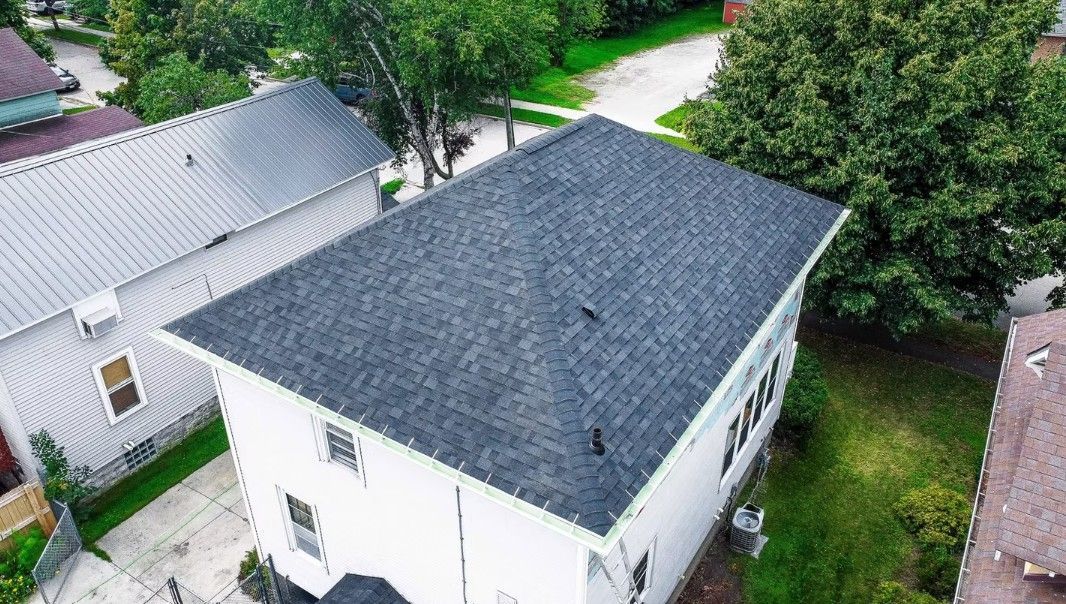 Aerial view of a white house with a dark shingled roof beside trees and neighboring homes.