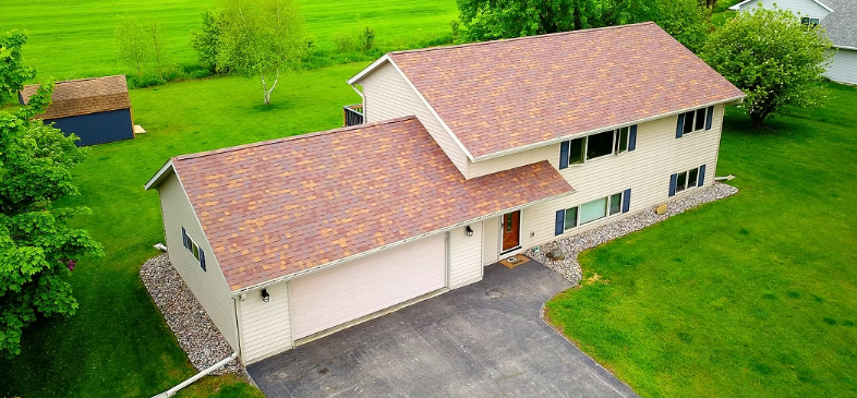 Aerial view of a beige two-story house with a red roof, garage, and driveway surrounded by green lawn.