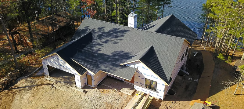 Aerial view of a large gray-roofed house beside a wooded lakeshore