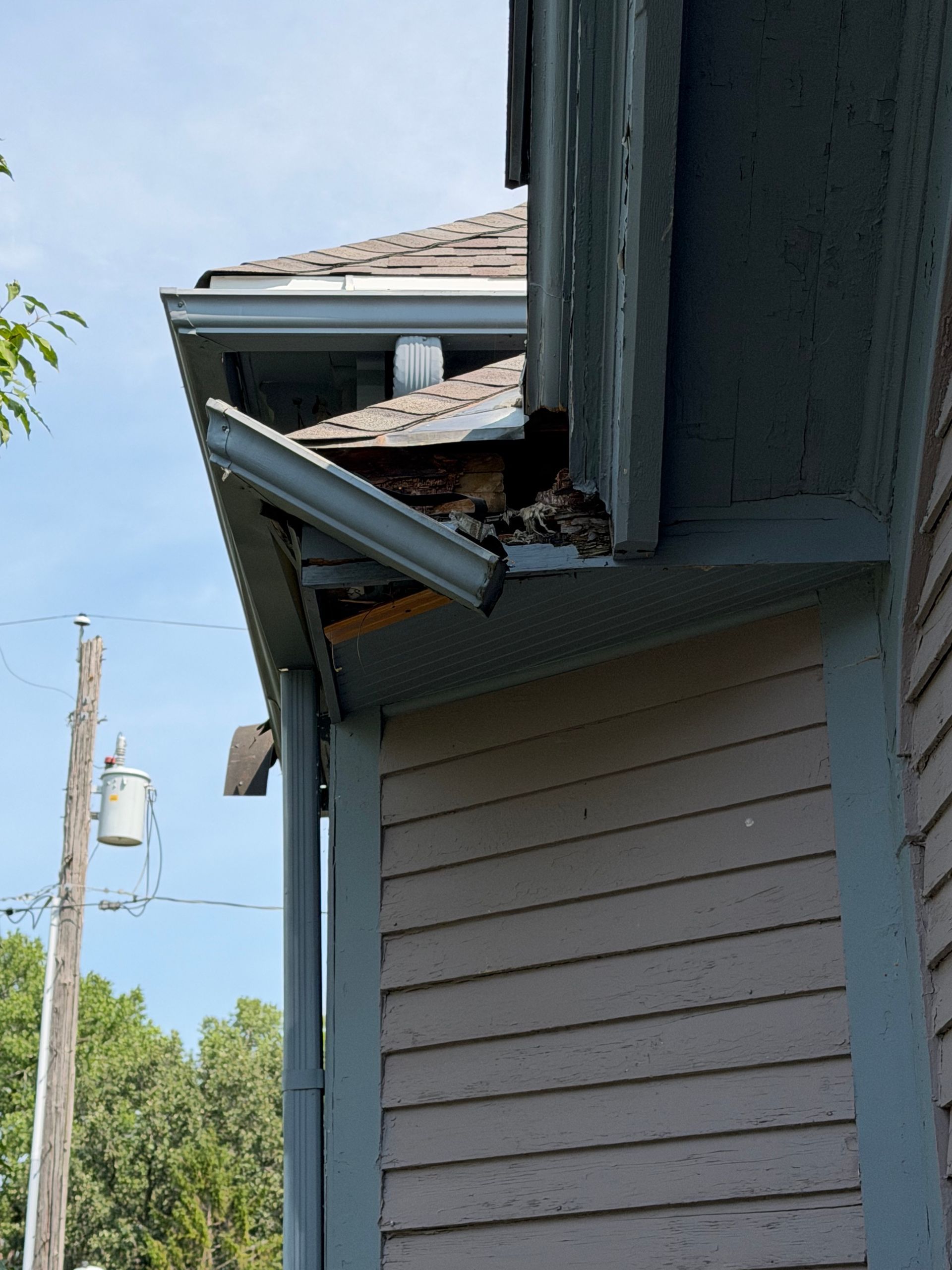 Damaged roof eaves and gutter on a gray house with broken siding, viewed from below.