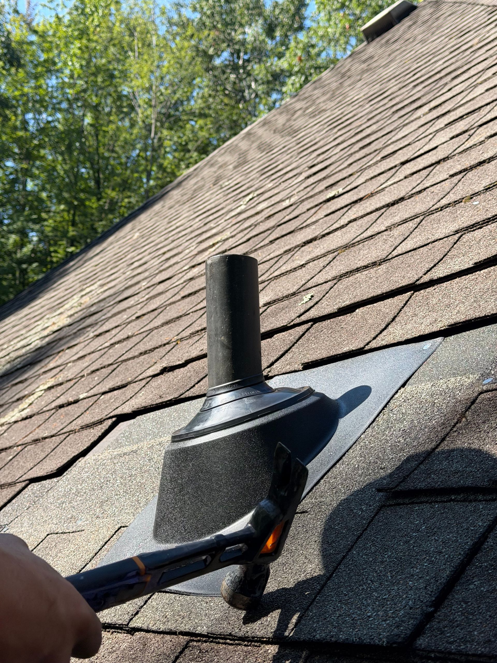 Hand tool scraping around a black roof vent on asphalt shingles under a blue sky