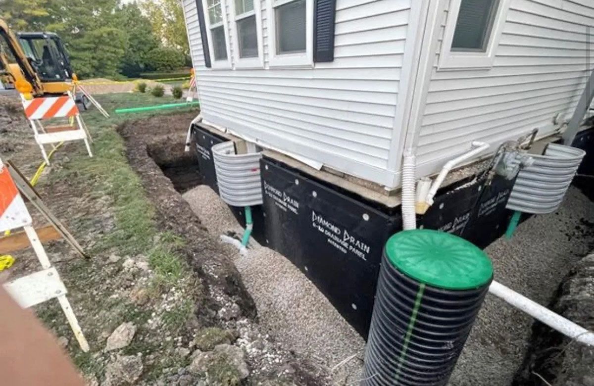 House foundation under excavation beside gray siding, with exposed black waterproofing and utility pipes
