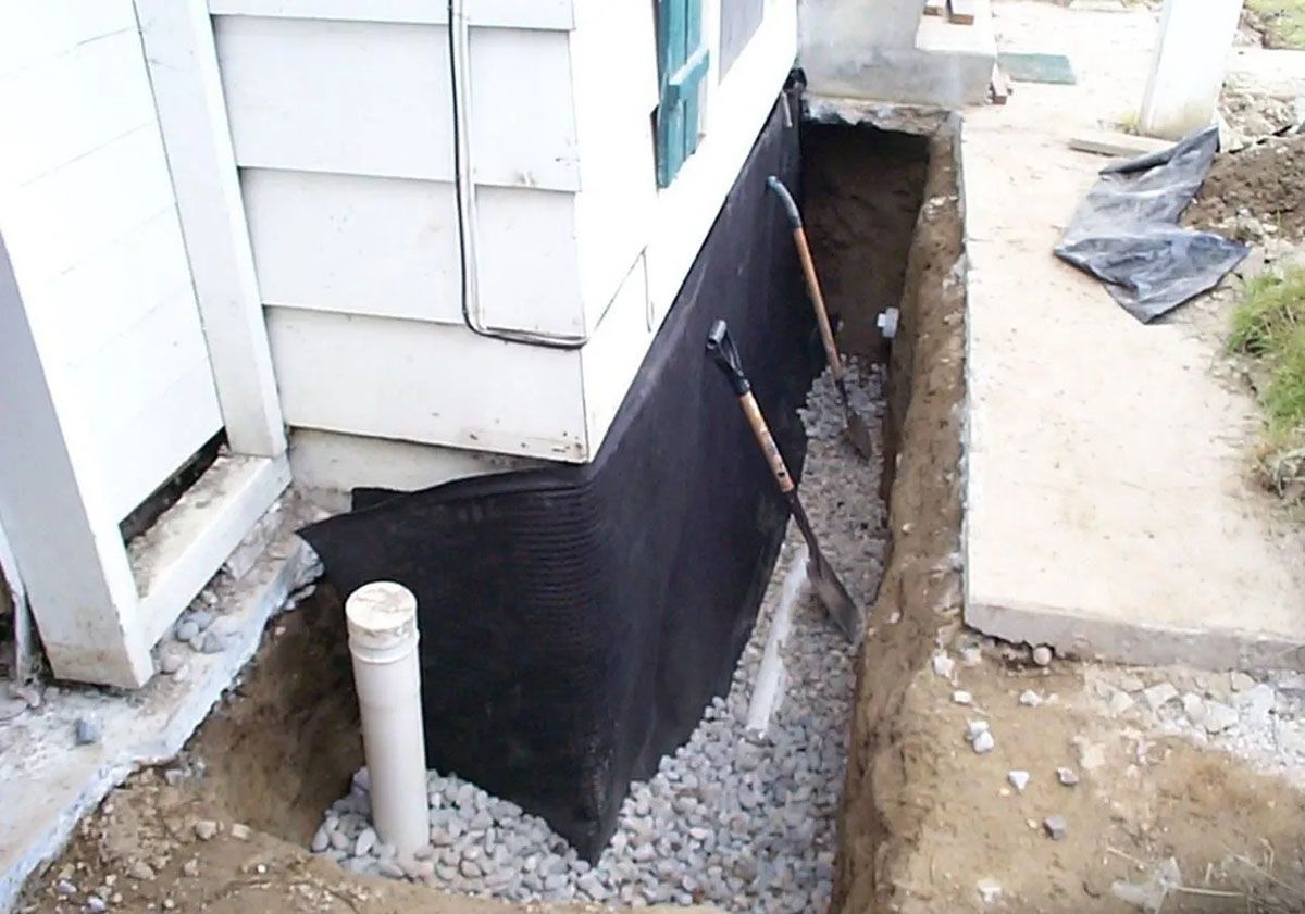 House foundation wrapped in black waterproofing with a trench and exposed pipe