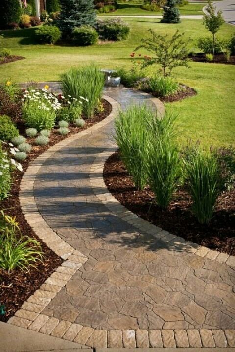 A curved stone pathway through a manicured garden with various plants and grasses, leading towards a small water feature.