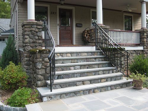 Stone steps lead up to a house with a brown door. Black iron handrails and stone pillars frame the entrance.