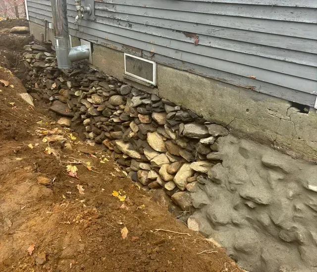 Rocks piled against a weathered house foundation. Dirt and trench visible. Gray siding and concrete.