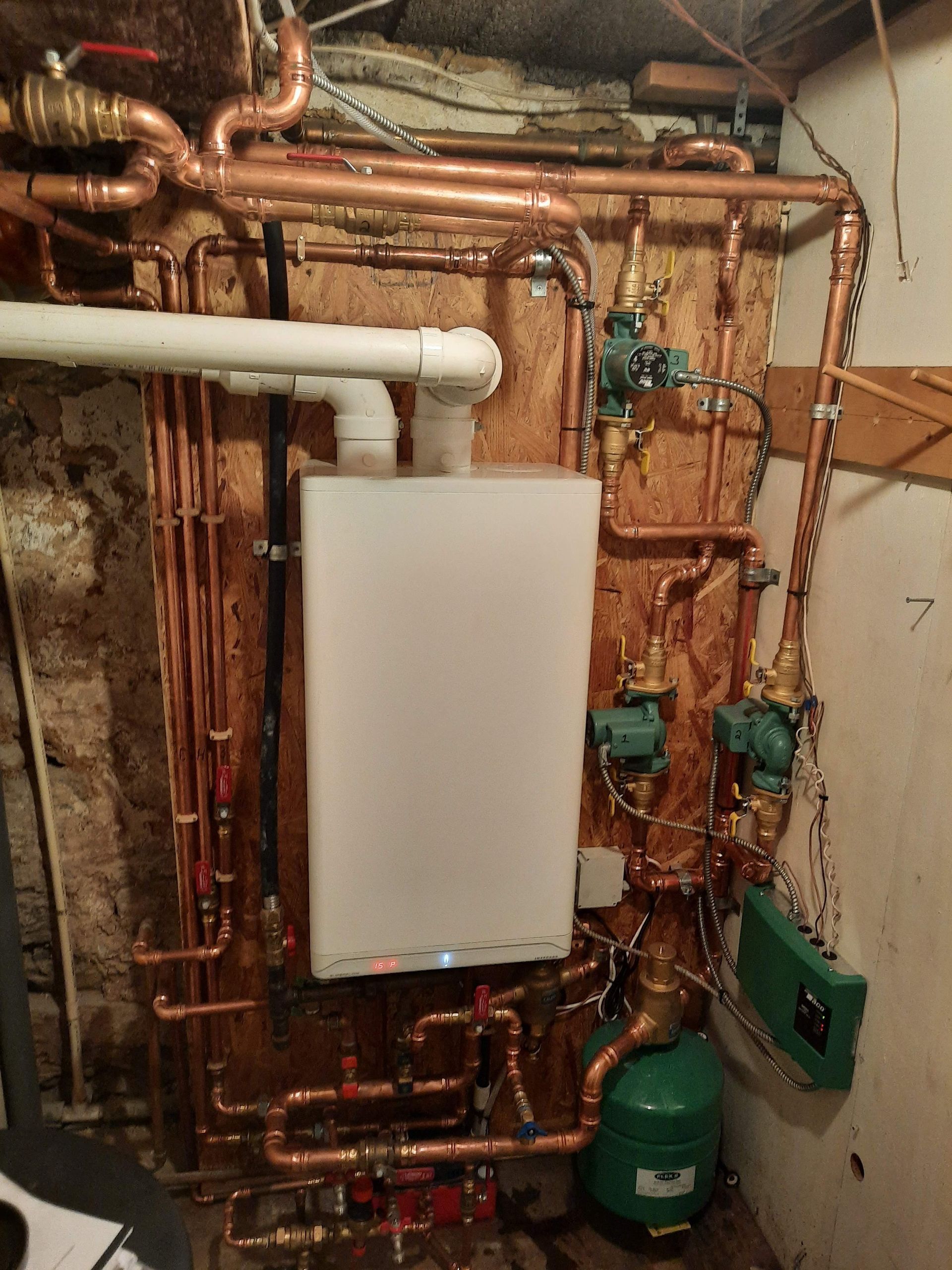 A white boiler is surrounded by copper pipes in a basement.