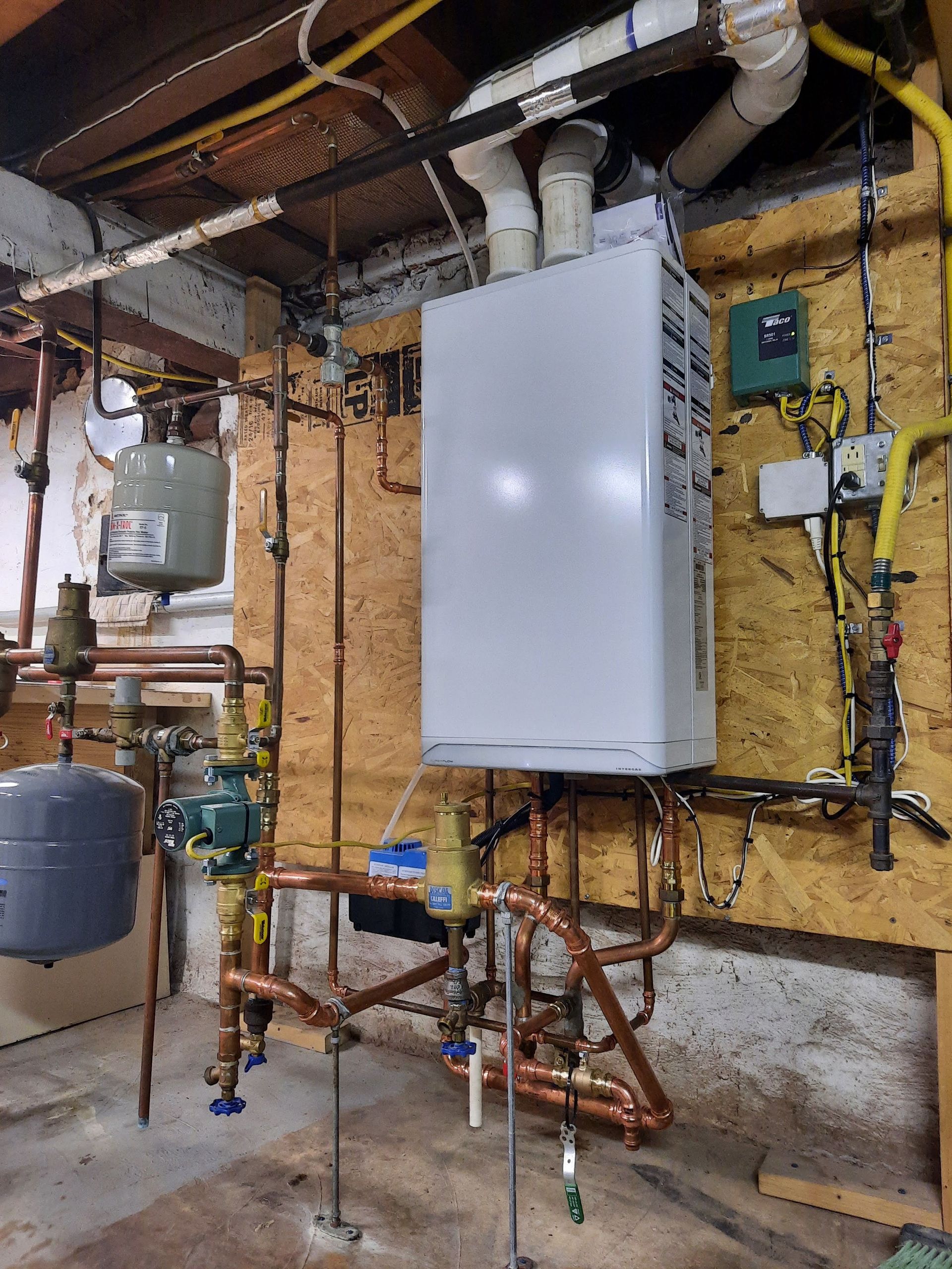 A boiler is sitting in a basement surrounded by pipes.