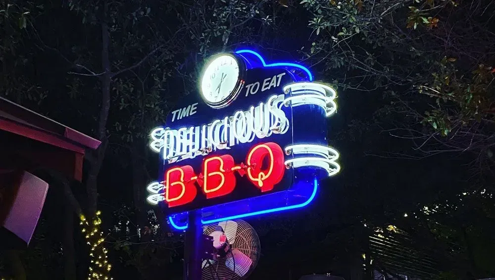 Neon sign for The Firehouse BBQ at night; blue, red, and white lights.