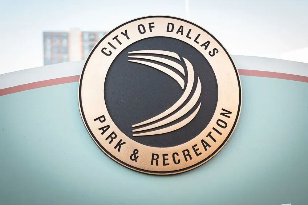 City of Dallas Park & Recreation logo, circular bronze sign on a light blue background.