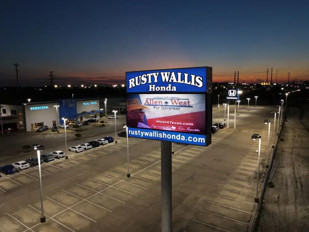 Sign for Rusty Wallis Honda at dusk, with lit parking lot and building in the background.