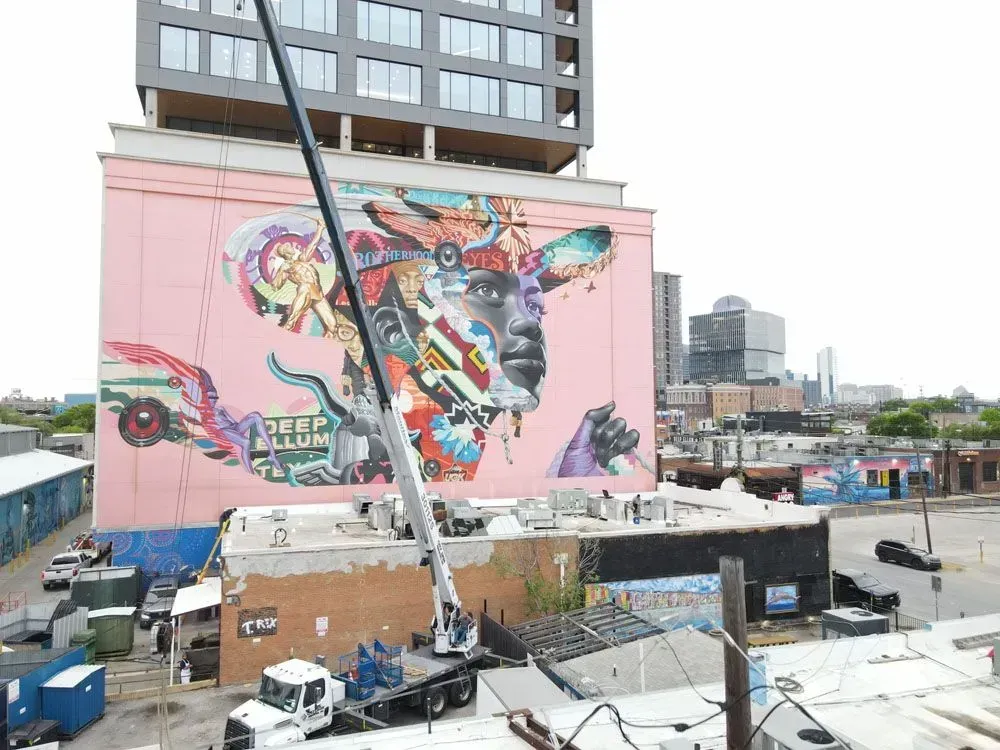 Crane operating near a large, colorful mural on a building. City setting with other buildings and vehicles.