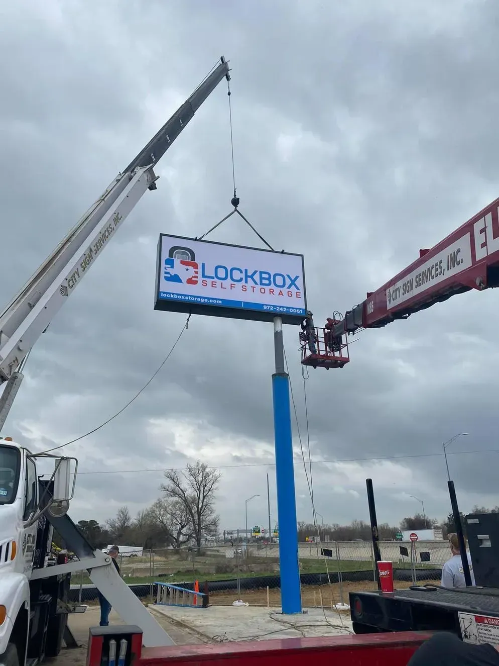 Two cranes installing a Lockbox Solutions sign on a blue pole. Workers in a lift. Overcast sky.
