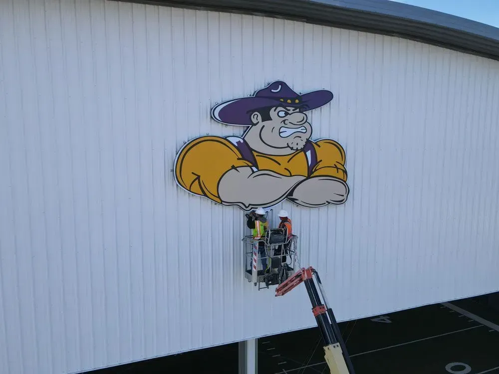 Workers in a lift installing a large cartoon mascot on a white building near a sports field.