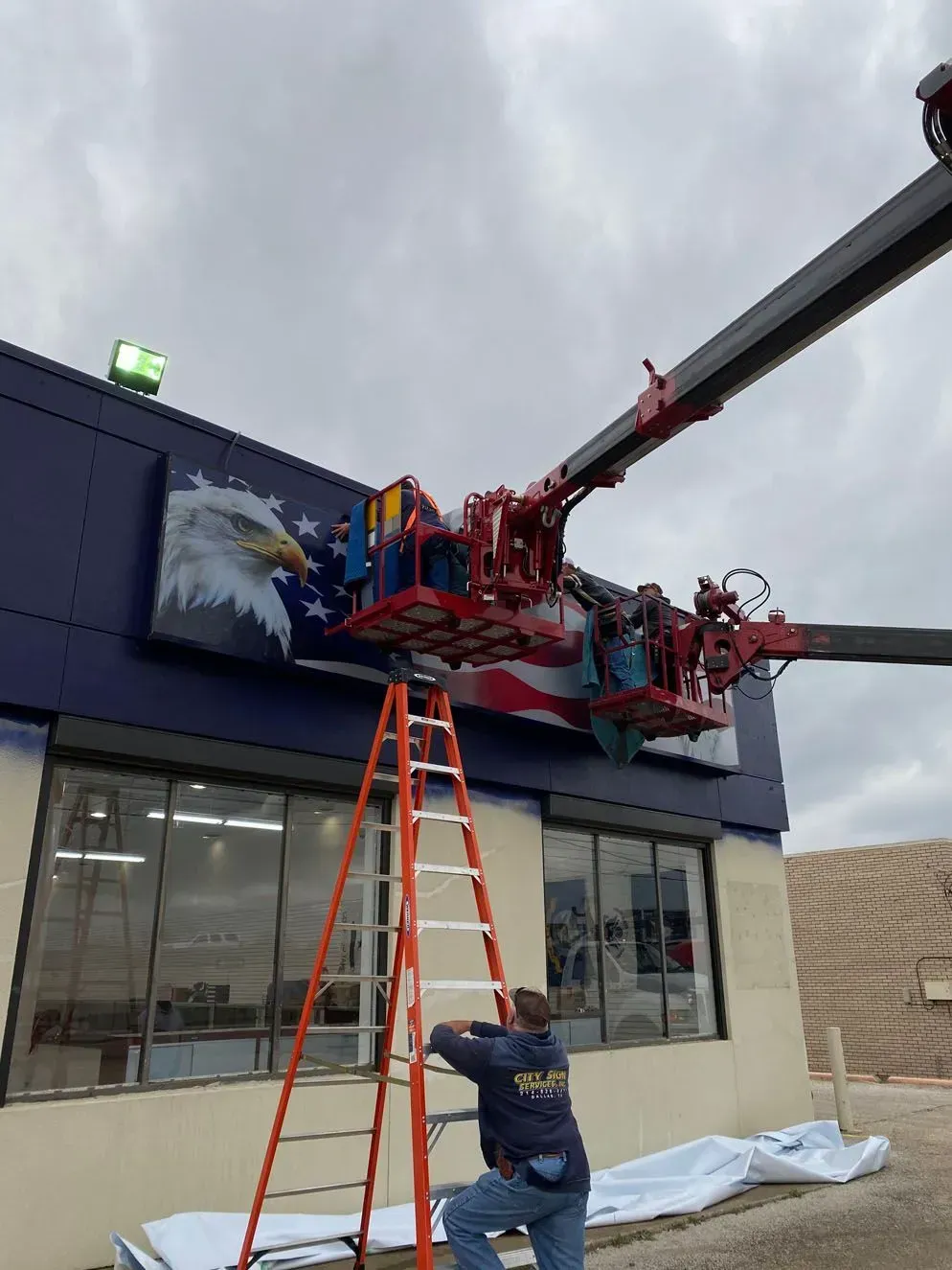 Person on a ladder, another in a lift painting a patriotic mural of an eagle and flag on a building.