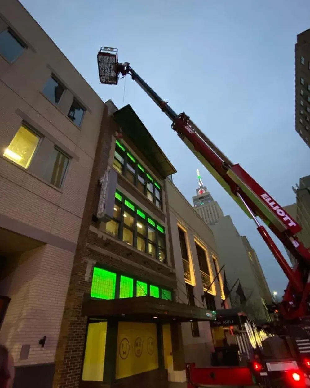 A crane with a bucket is working on a building with green neon lights. The sky is twilight.