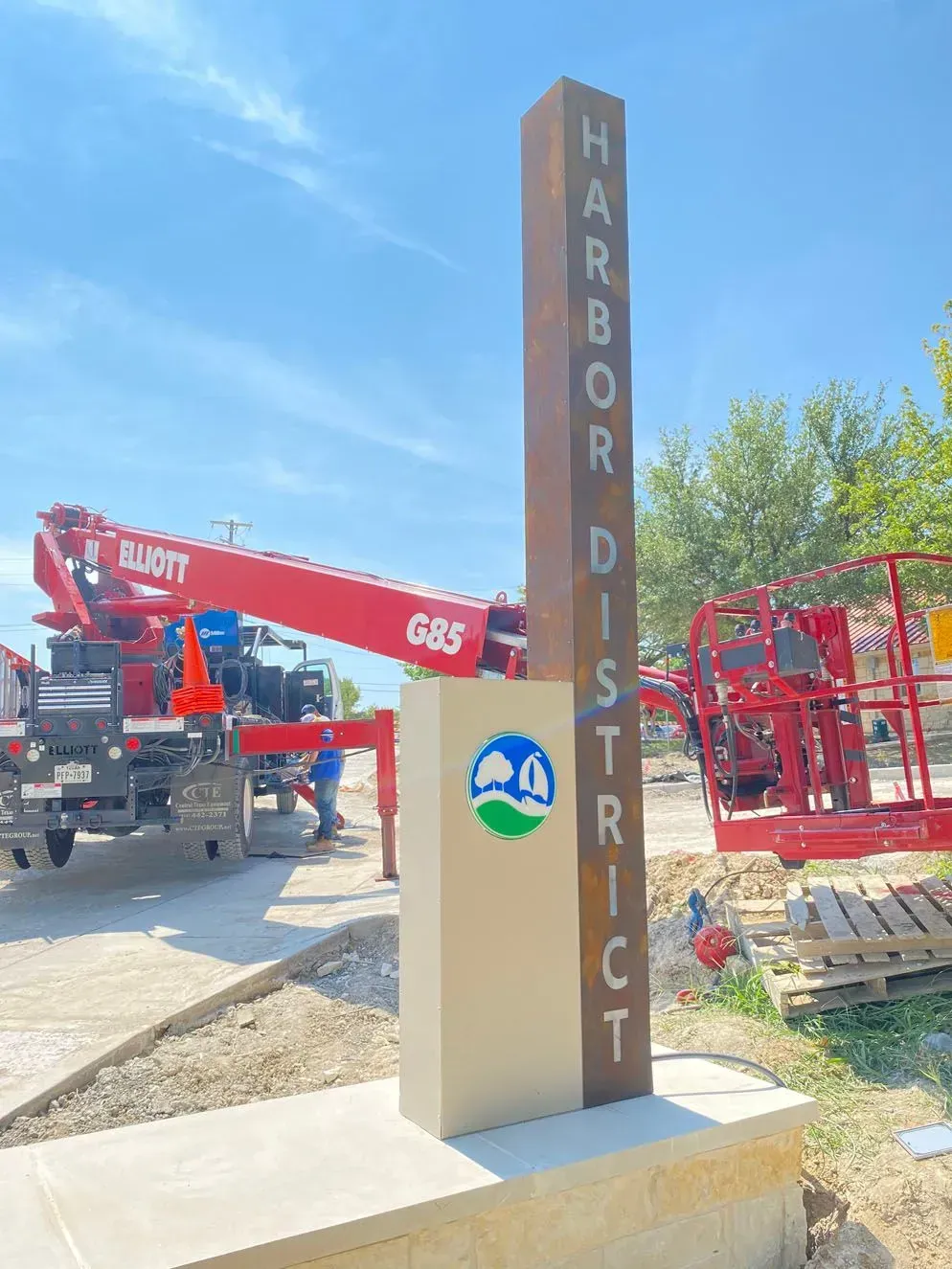 Sign for Harbor District. Red crane, blue sky, concrete base, and logo with blue and green.