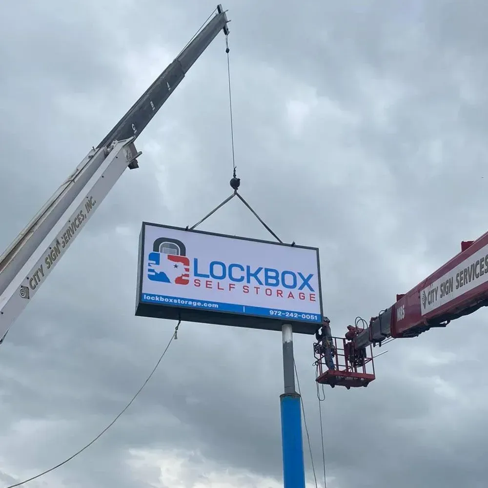 A sign for Lockbox Self Storage being installed by a crane and workers in a lift.