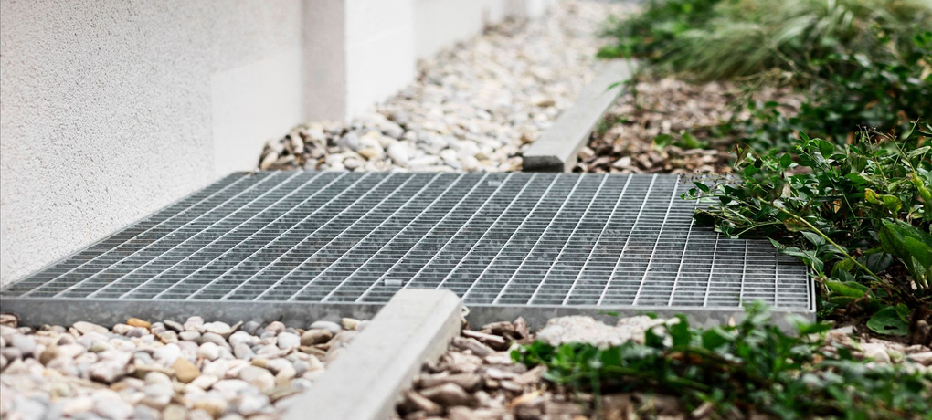 Metal grate over pebbles and landscaping, next to a building wall.