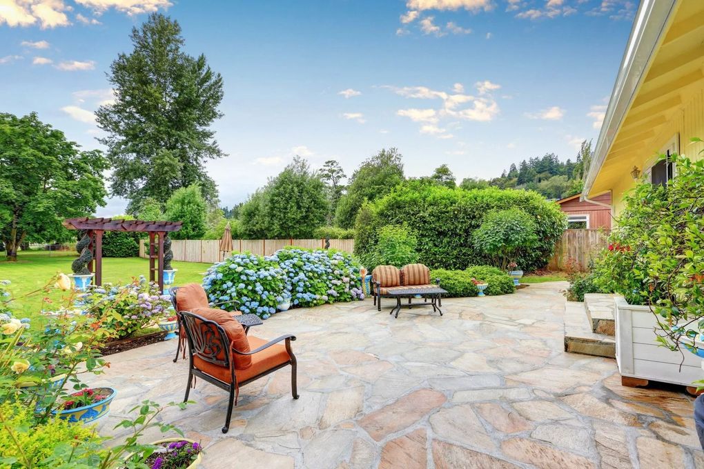 Patio with stone pavers, chairs, and hydrangeas against a lush green backdrop, under a blue sky.