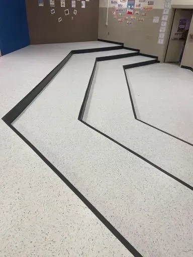 School hallway with white floor and black taped lines forming a geometric path.