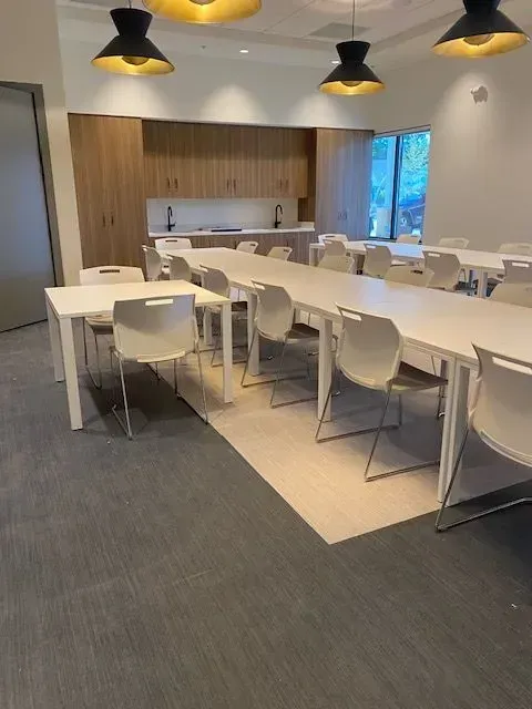 Modern office break room with white tables and chairs, wood cabinets, gray carpet, and black pendant lights