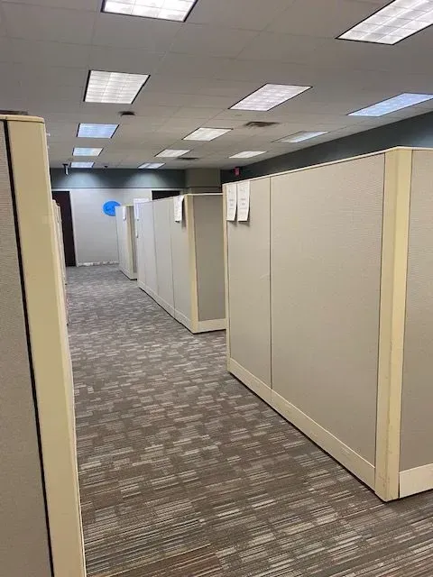 Empty office hallway with beige cubicle walls, gray carpet, and fluorescent ceiling lights.