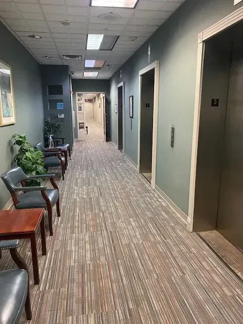Office hallway with carpet, chairs, potted plants, and elevator doors at the end