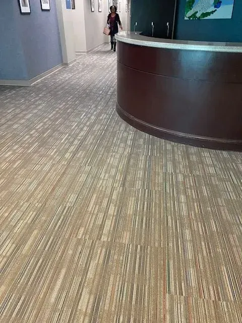 Office hallway with gray carpet and a curved dark wood reception desk; a person walks in the distance.