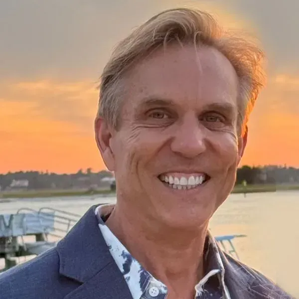 Smiling man in a blue blazer at sunset near a waterfront.