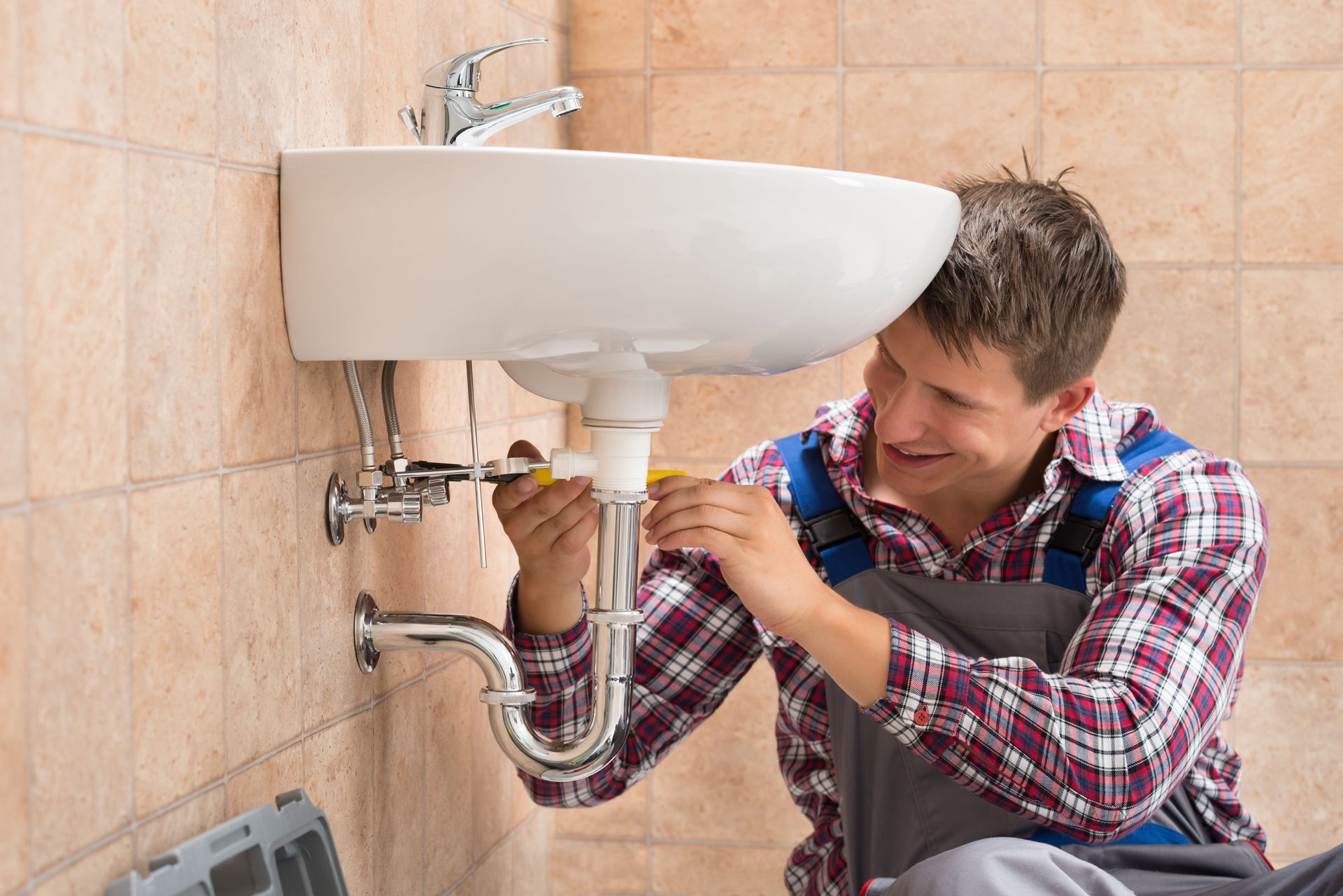 A smiling person in work clothes repairing plumbing pipes under a white bathroom sink against a tiled wall.