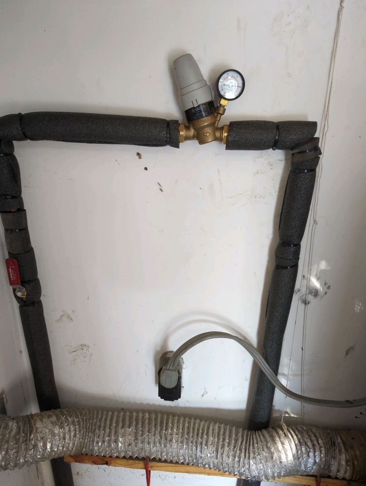 Wall-mounted plumbing pipes with black insulation and a pressure gauge in the center.