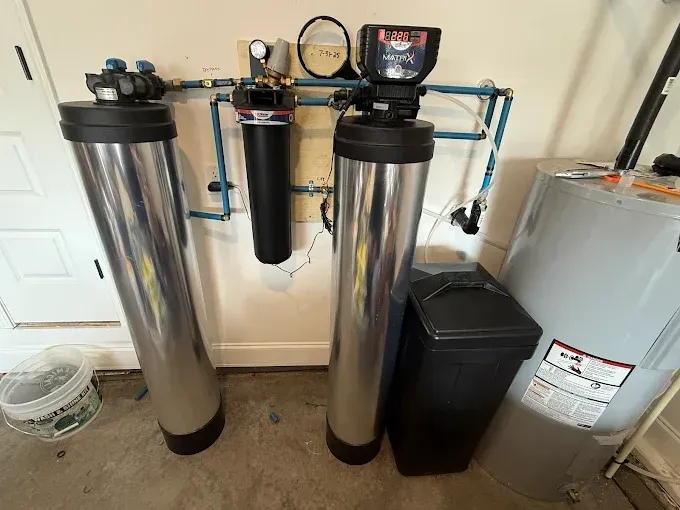 Whole-house water filtration system with two tall stainless tanks, wall-mounted filters, and a salt tank in a basement.
