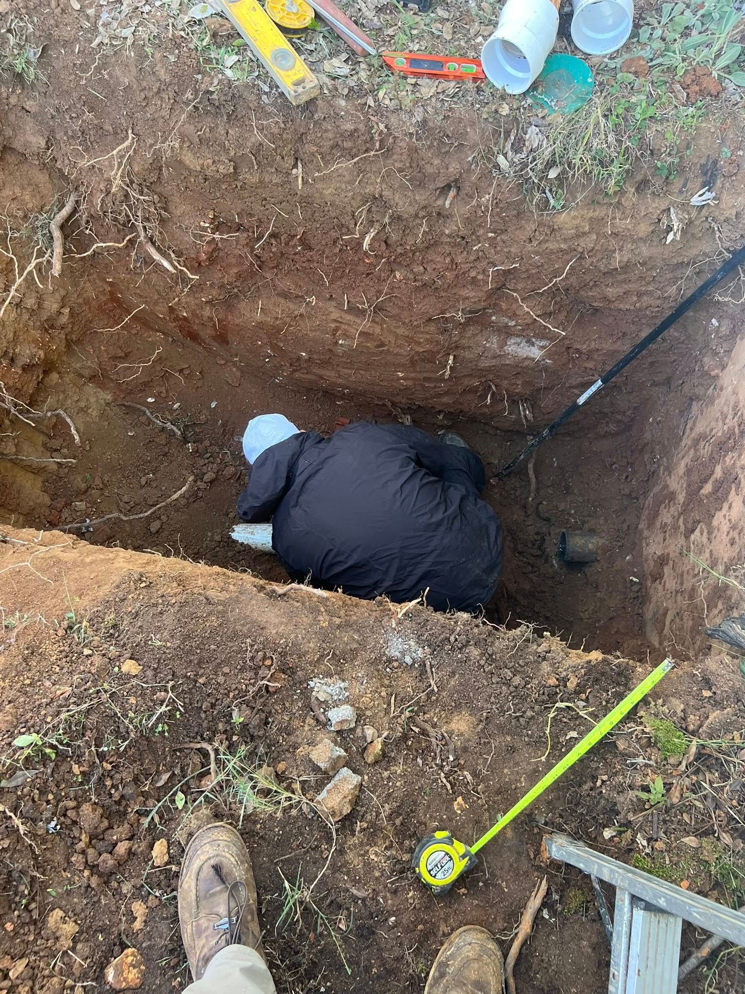 Worker in a deep dirt trench, wearing dark clothing, with tools and hoses around the edge.