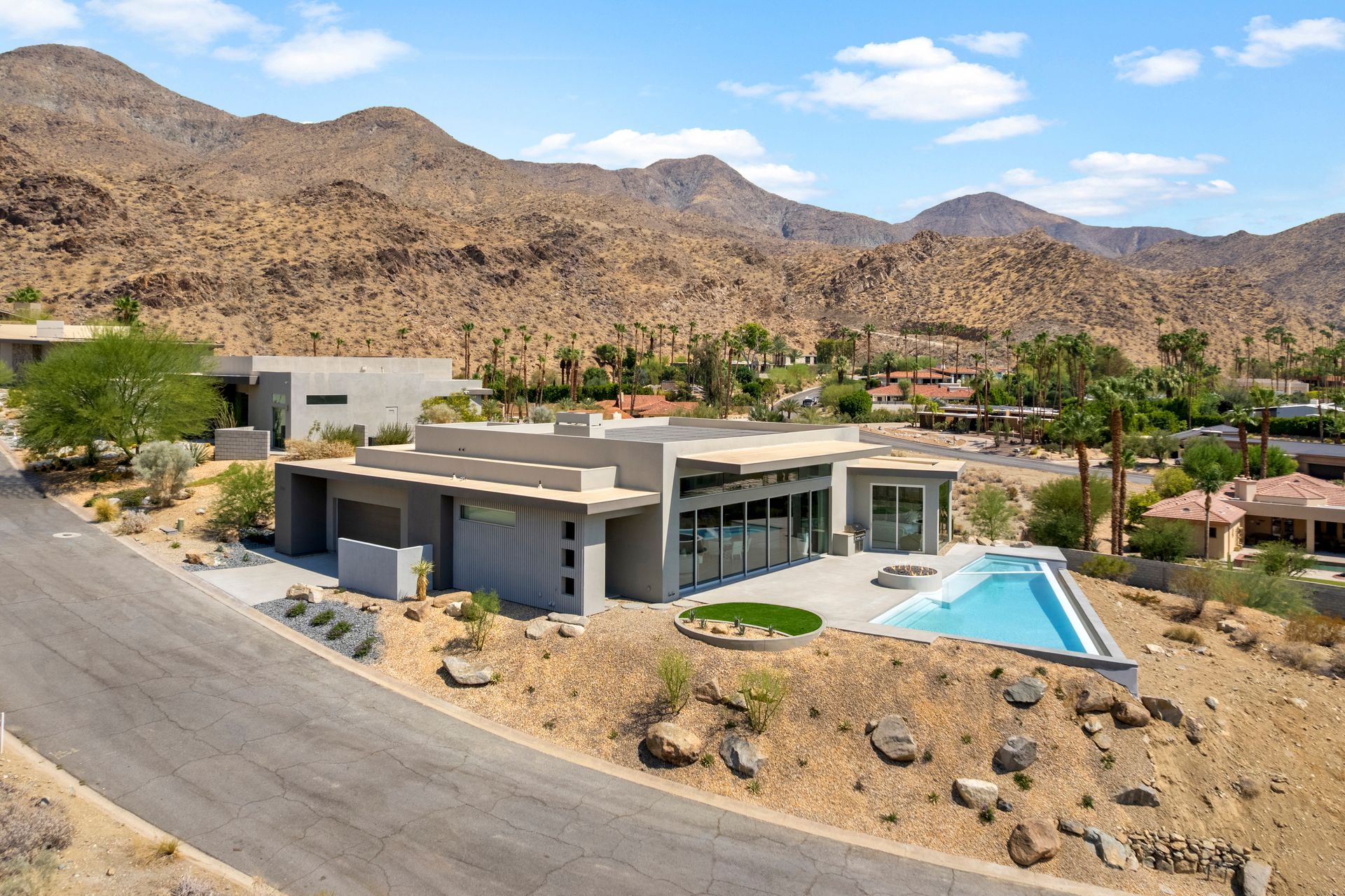 A modern, gray-toned flat-roof house with a rectangular swimming pool, set in a desert landscape against rocky mountains.