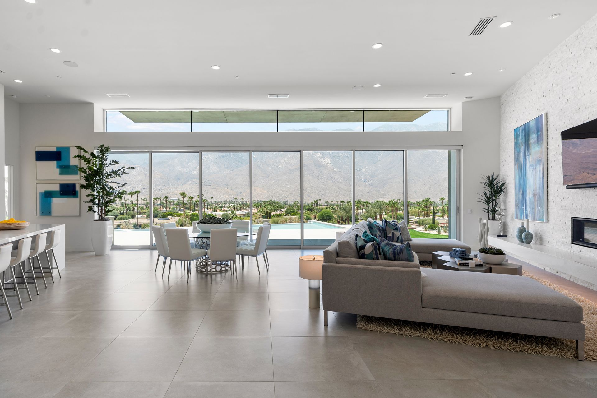 A modern, bright open-plan living room with a large gray sectional, dining set, and floor-to-ceiling mountain views.