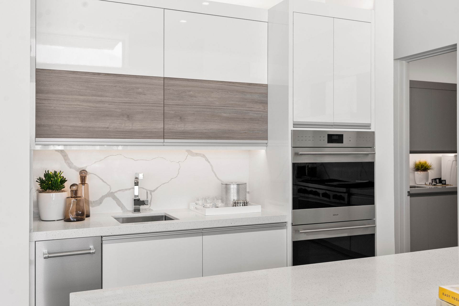 Modern kitchen with white glossy cabinets, wood-grain accents, a marble backsplash, and a built-in stainless steel oven.