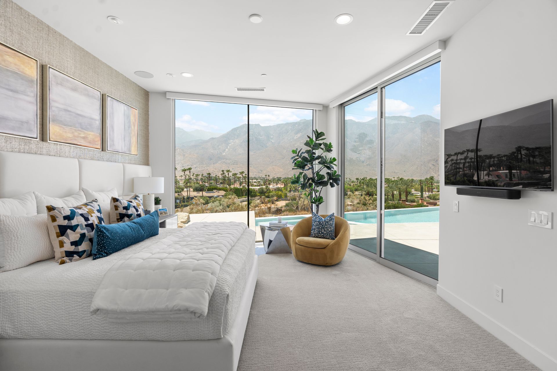 Modern bedroom with white decor, a large bed, a gold accent chair, and a floor-to-ceiling view of mountains and a pool.