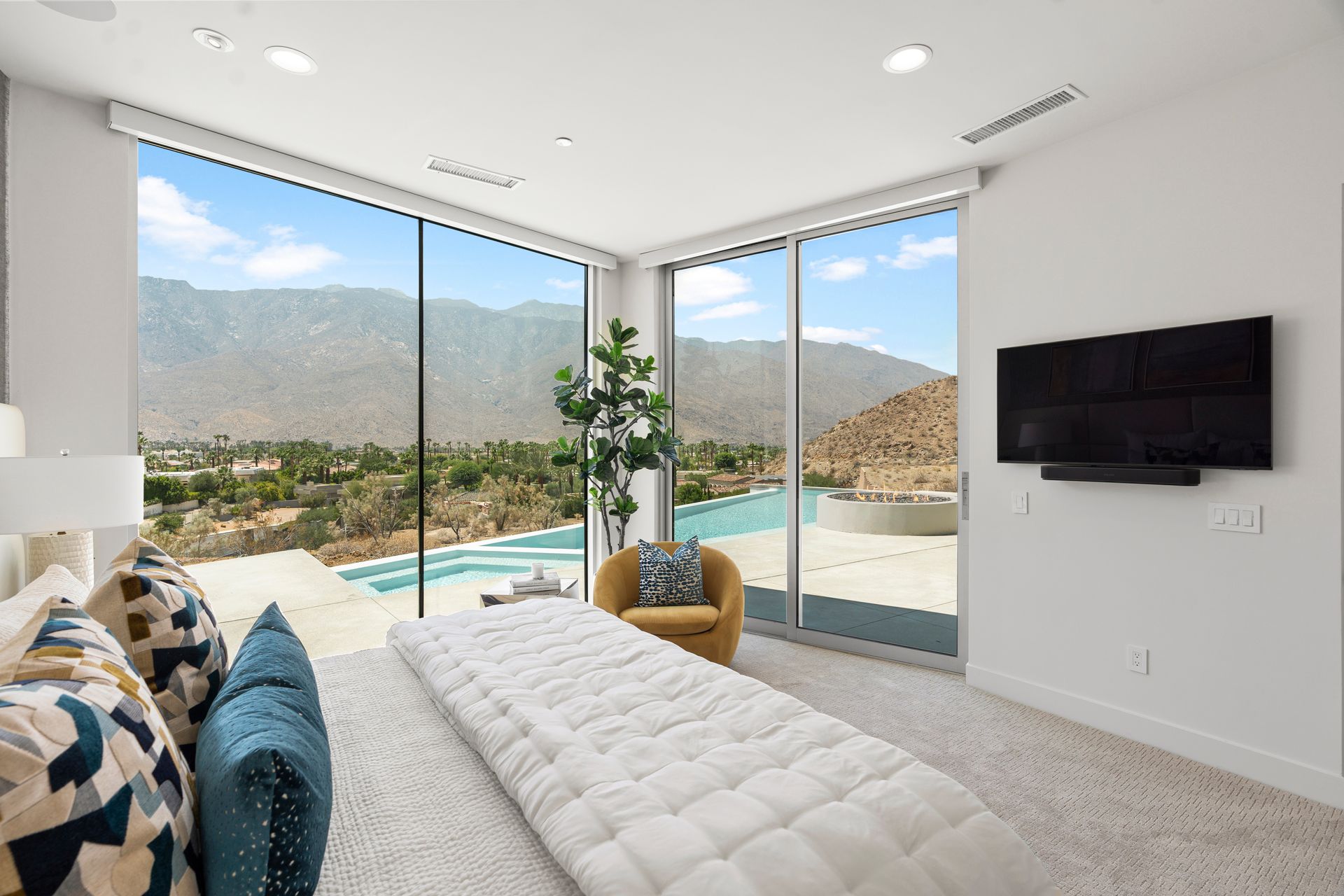 A modern bedroom with a large bed, a gold armchair, and floor-to-ceiling windows overlooking a pool and desert mountains.