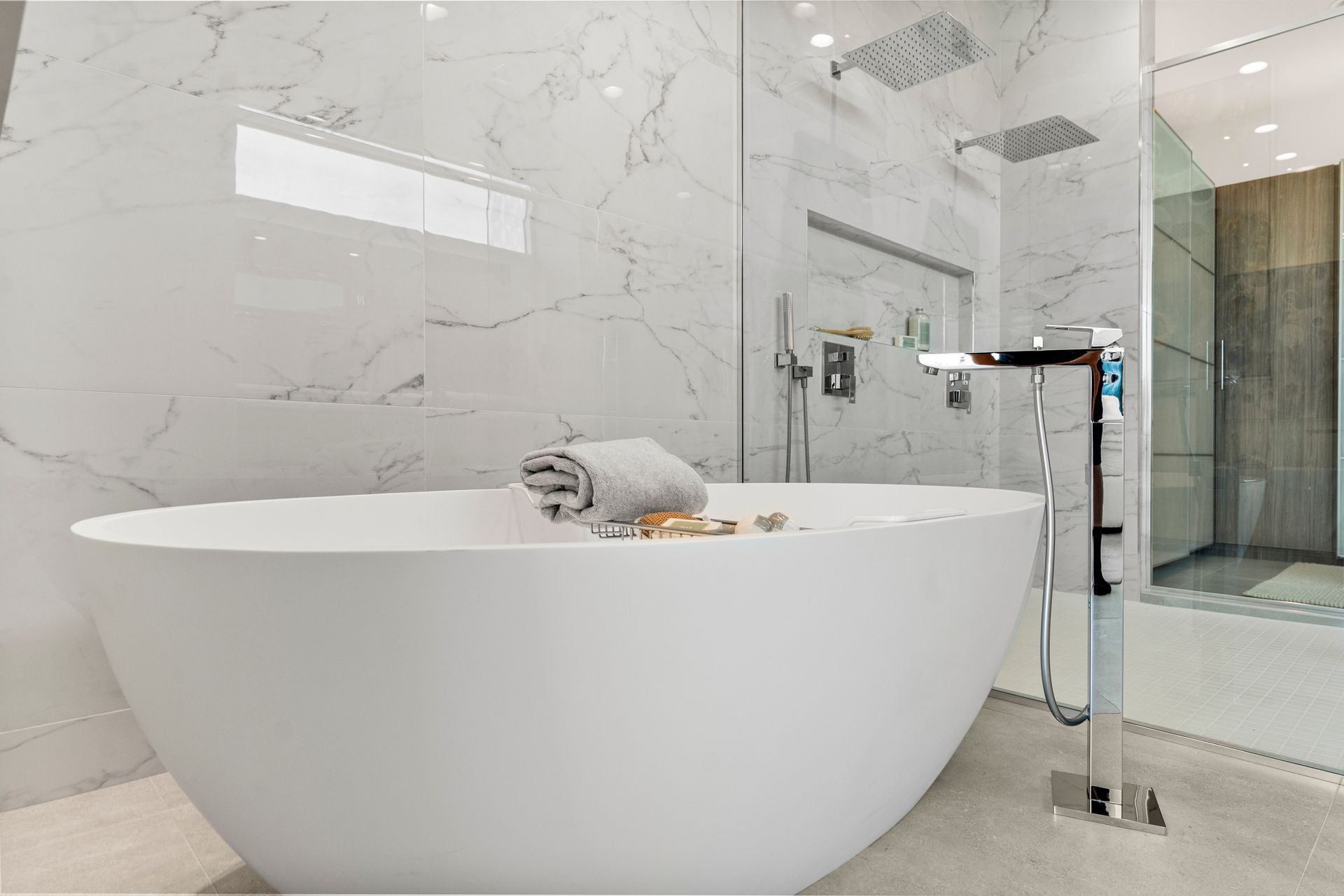 A white, oval freestanding bathtub sits in a modern bathroom with marble-tiled walls and a glass-enclosed shower.