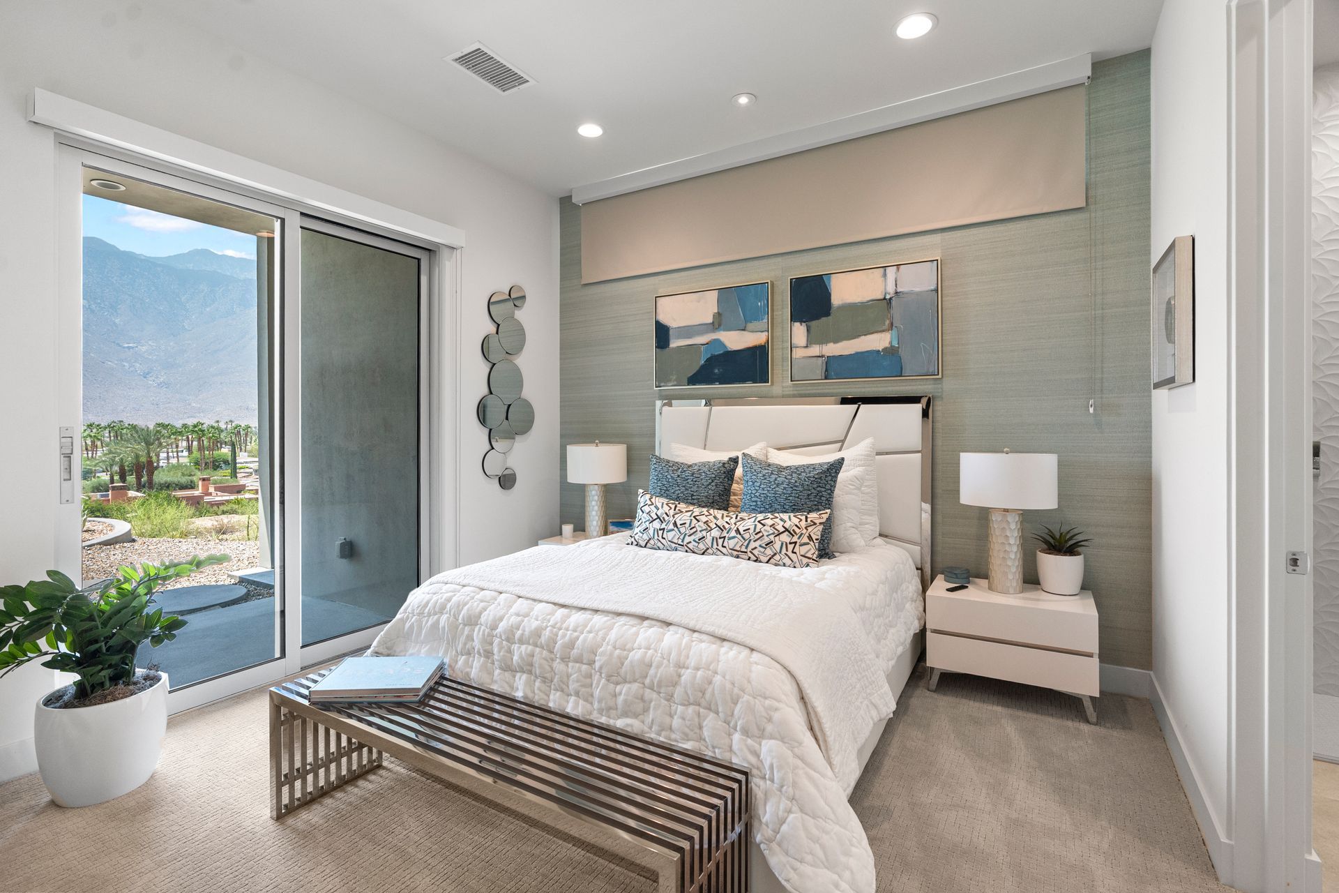 A modern bedroom featuring a bed with white bedding, blue pillows, two nightstands, and a glass door with a mountain view.