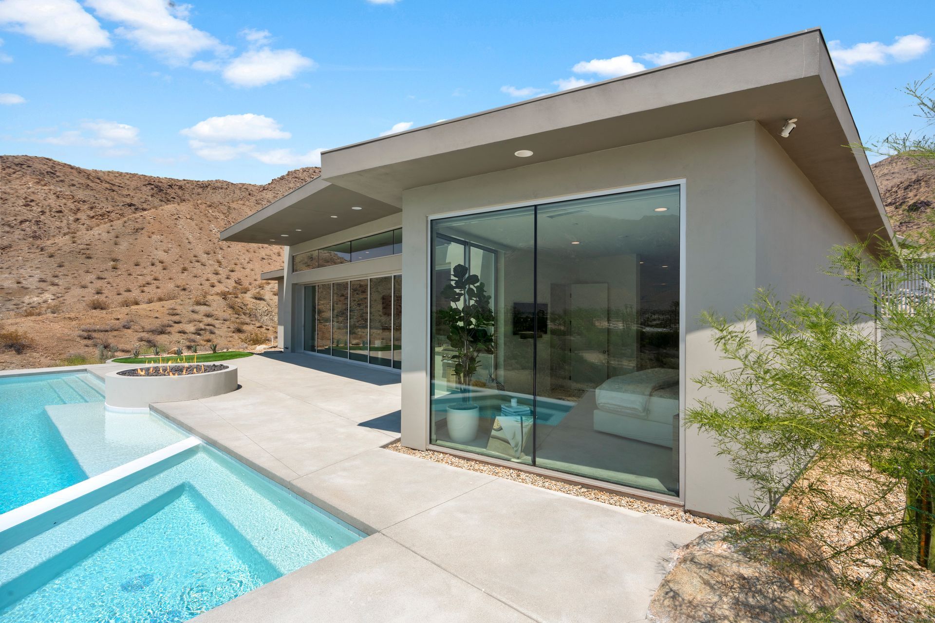 Modern house with floor-to-ceiling glass walls, a blue swimming pool, and a desert mountain landscape in the background.