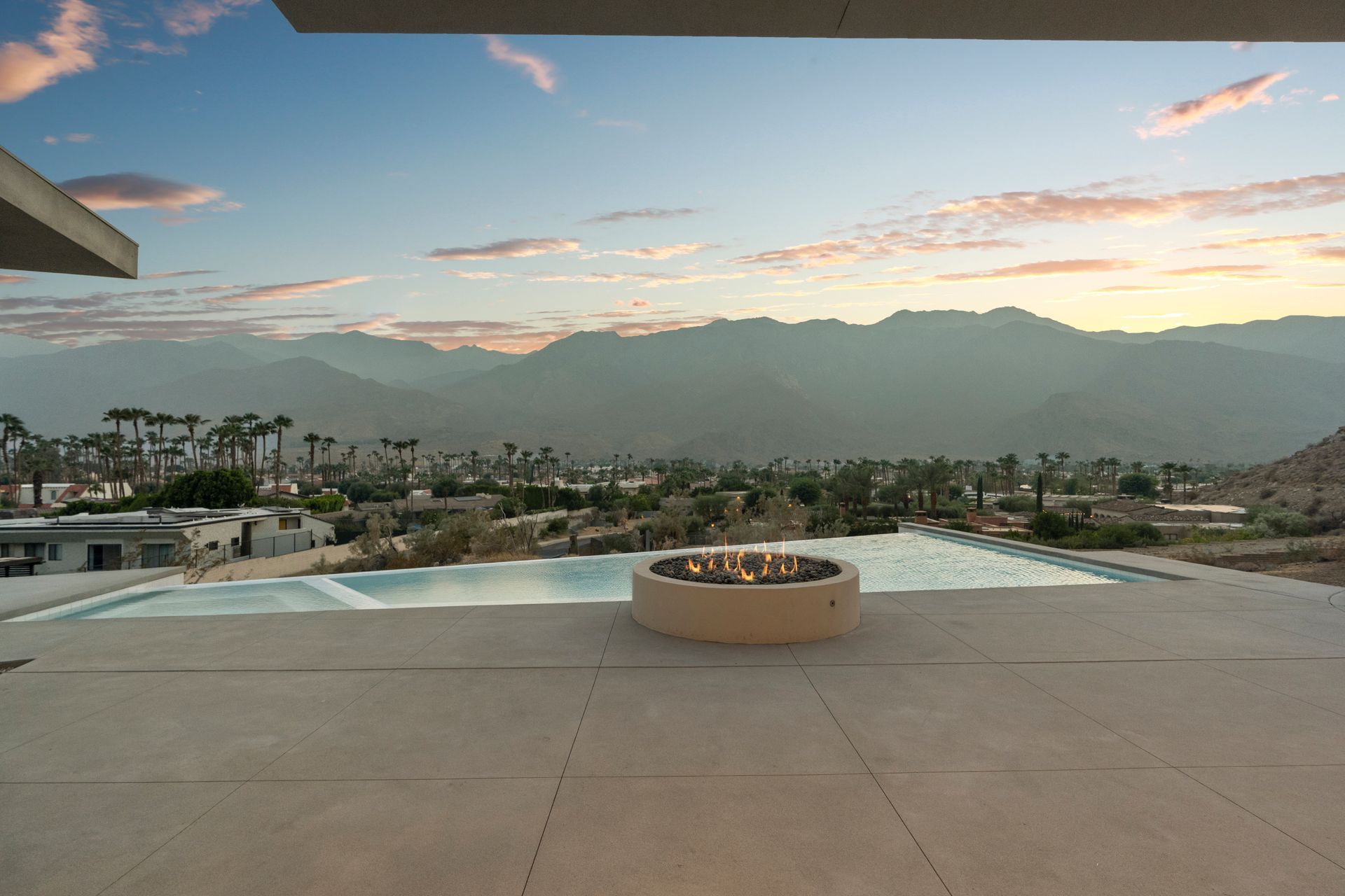 A modern concrete patio with a circular fire pit overlooks an infinity pool and valley against a desert mountain sunset.