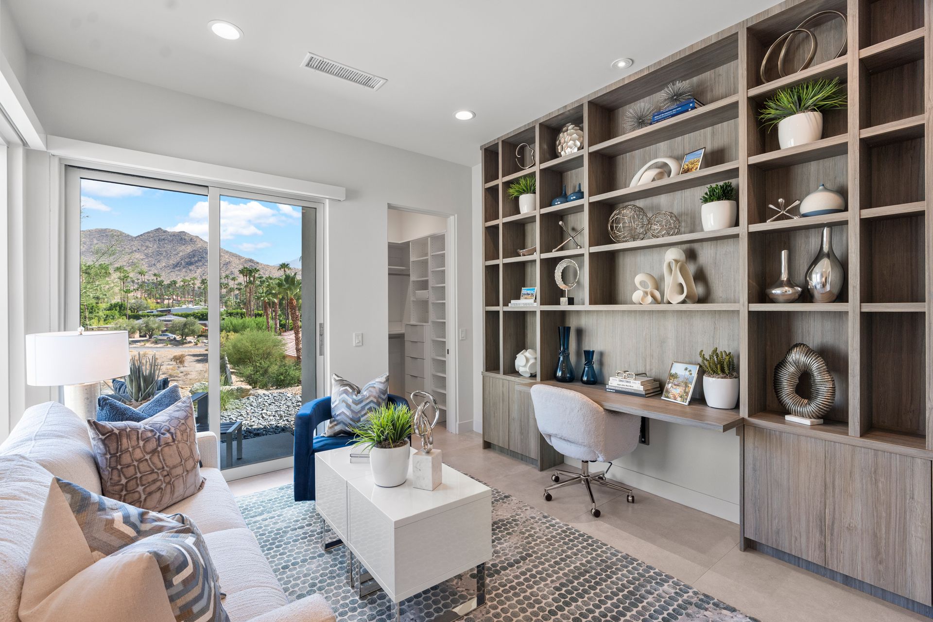 A home office with built-in wooden shelves, a desk, a white rolling chair, a sofa, and a scenic desert mountain view.