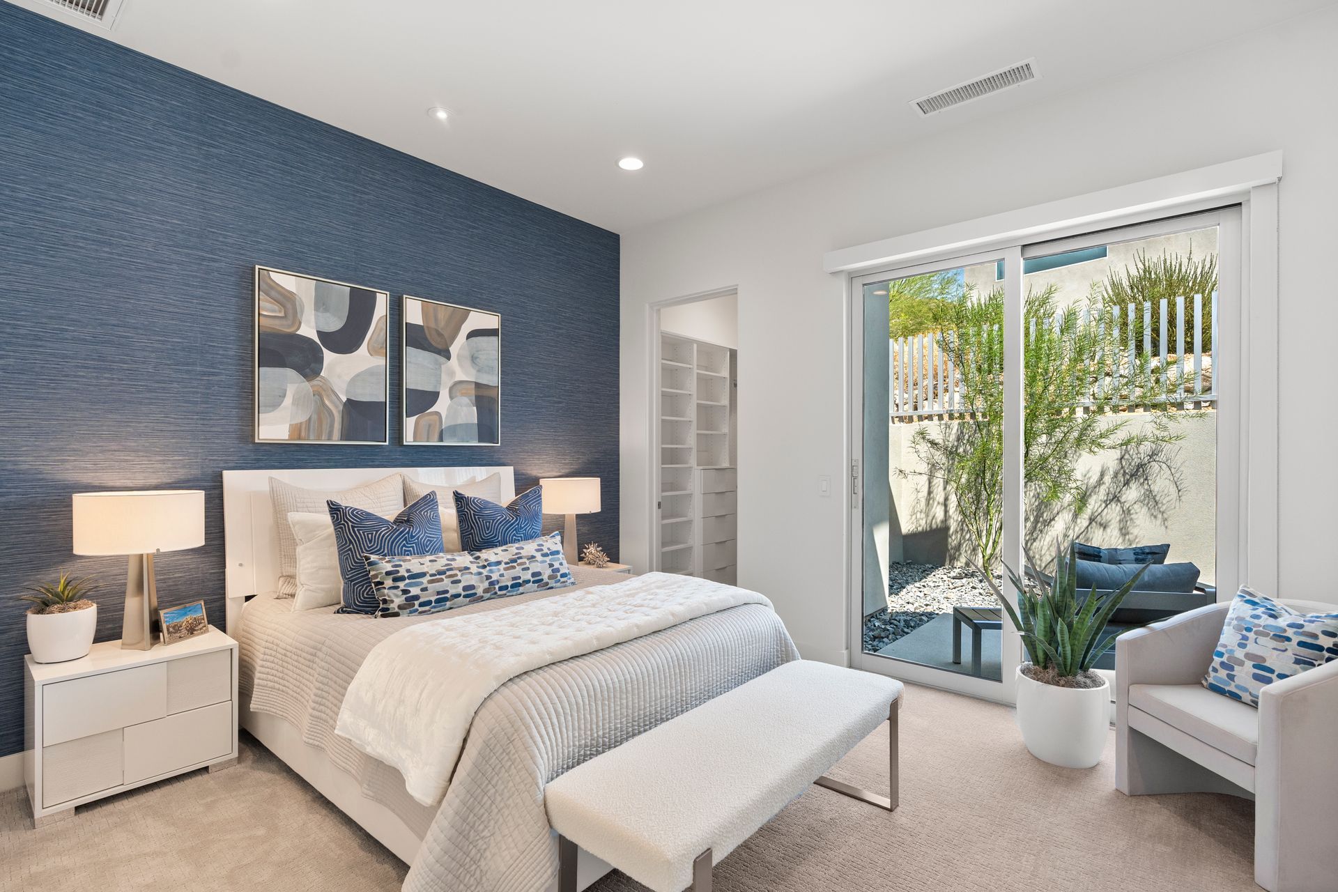 A bedroom with a blue textured accent wall, a white bed, a bench, a nightstand, and a glass door opening to a patio.