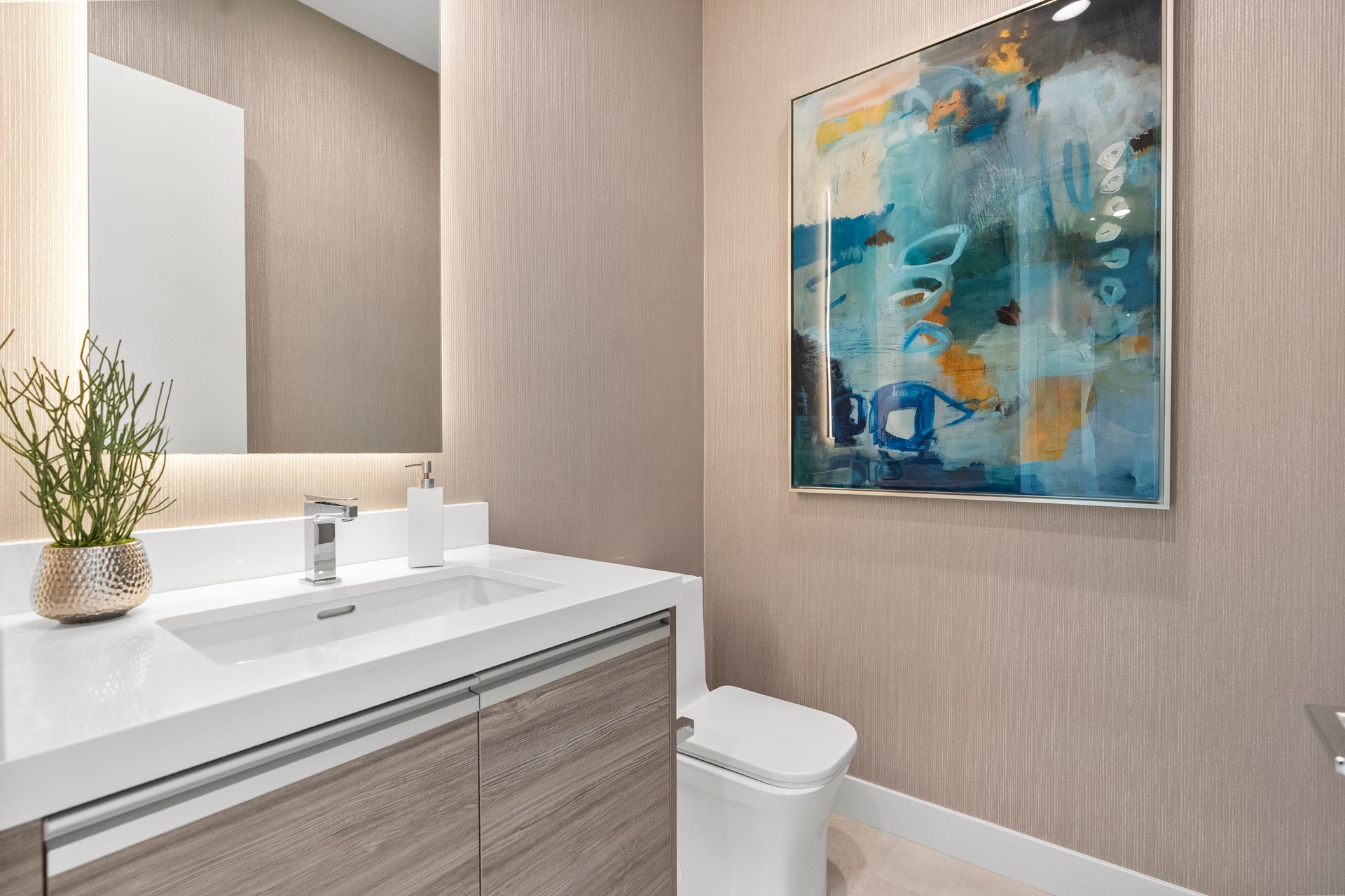 A modern bathroom with a wood-grain vanity, white countertop, a mirror, a potted plant, and abstract art on the wall.